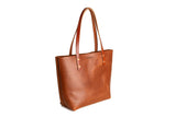 A brown leather tote bag with long handles displays on a white background.
