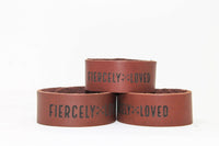Three brown leather bracelets displaying the text "Fiercely Loved" stack against a white background.
