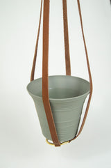 Hanging planter displays a sage green pot held by brown leather straps with a gold ring detail.

