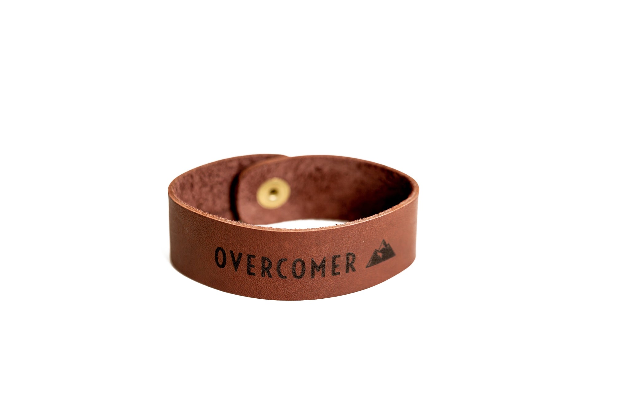 A brown leather bracelet displaying the word "OVERCOMER" and a mountain graphic rests on a white surface.
