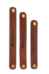 Three brown leather straps with brass buttons, each stamped with "OVERCOMER" and a mountain logo, lie vertically against a white background.
