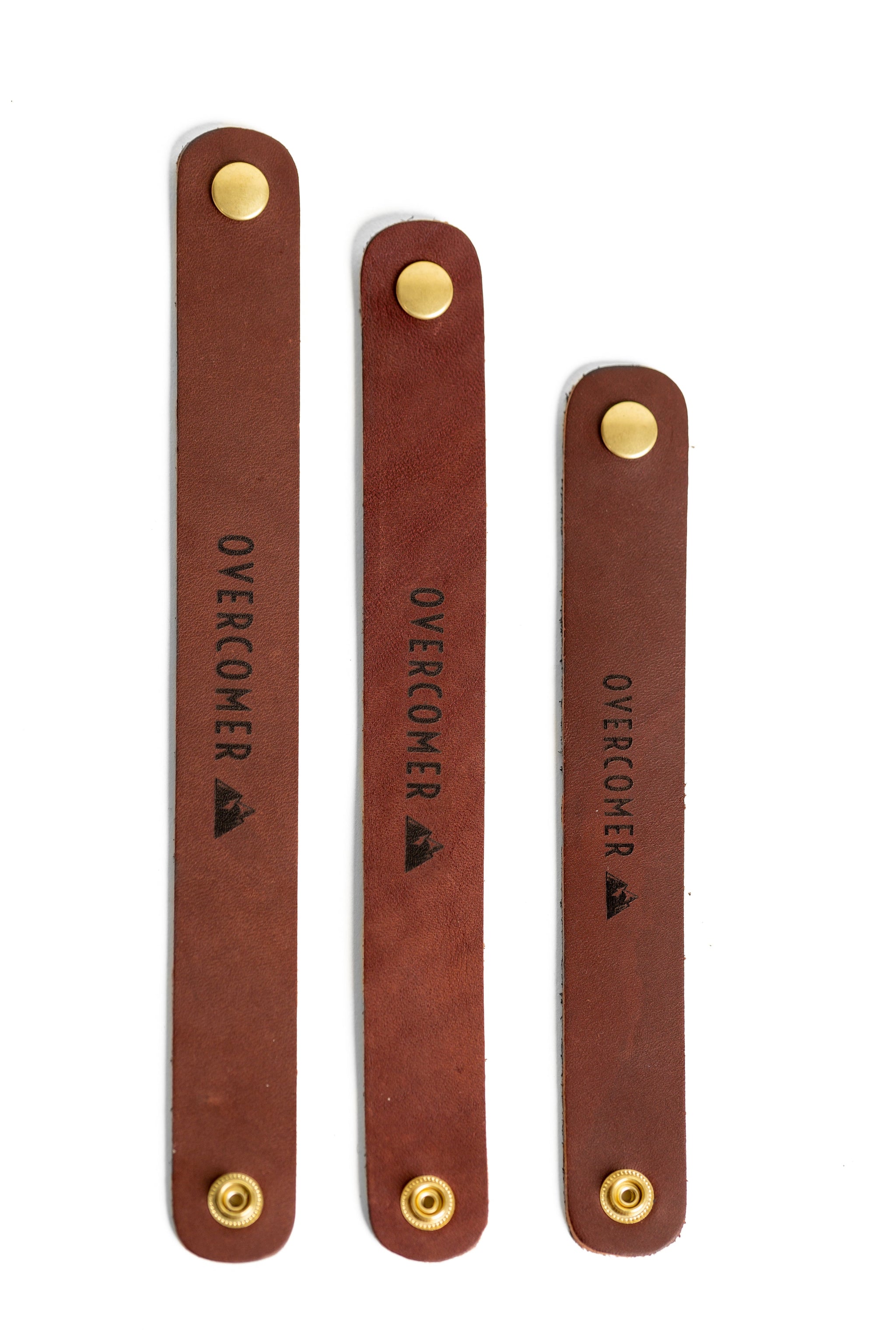 Three brown leather straps with brass buttons, each stamped with "OVERCOMER" and a mountain logo, lie vertically against a white background.
