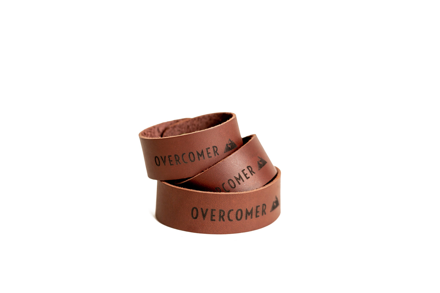 Three stacked brown leather bracelets, each stamped with "OVERCOMER" and a mountain logo, signify strength and resilience.
