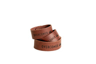 Three stacked brown leather bracelets, each stamped with "OVERCOMER" and a mountain logo, signify strength and resilience.
