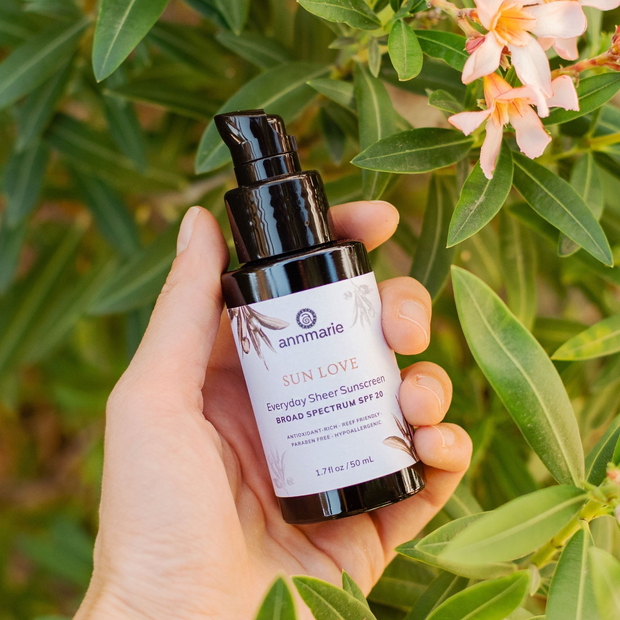 A hand holds Annmarie Sun Love sheer sunscreen SPF 20 in front of a leafy green plant with peach-colored flowers.
