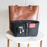 A brown leather tote bag sits behind a black felt purse organizer filled with a red smartphone, pen, and earbuds.
