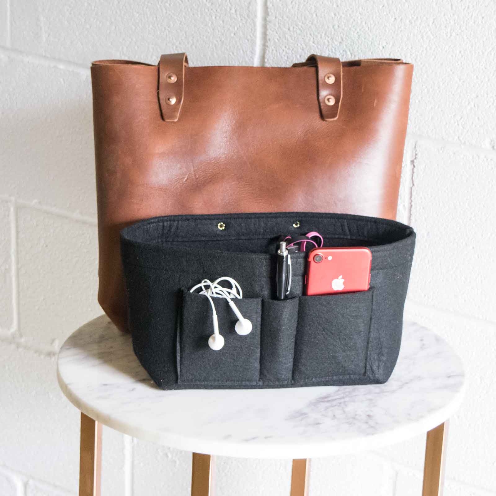 A brown leather tote bag sits behind a black felt purse organizer filled with a red smartphone, pen, and earbuds.
