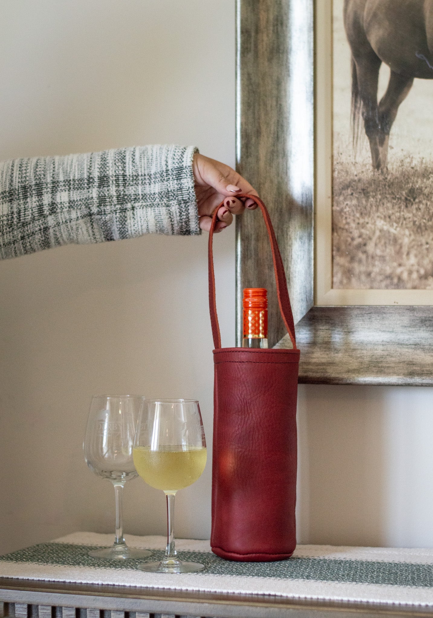 A hand lifts a red leather wine tote bag containing a wine bottle, next to wine glasses, for a stylish wine presentation.
